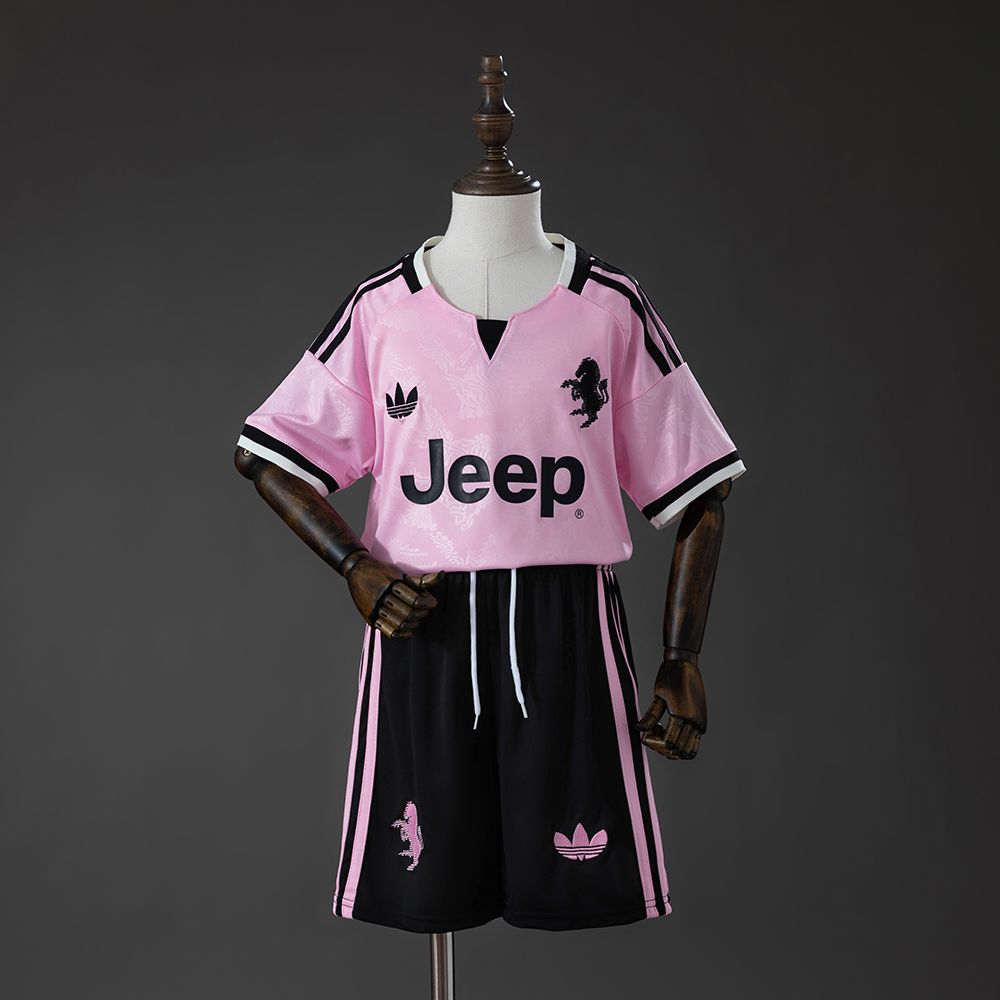 Rosa Juventus bortedrakt for barn med svarte shorts og rosa sidedetaljer, vist som komplett sett på dukke
